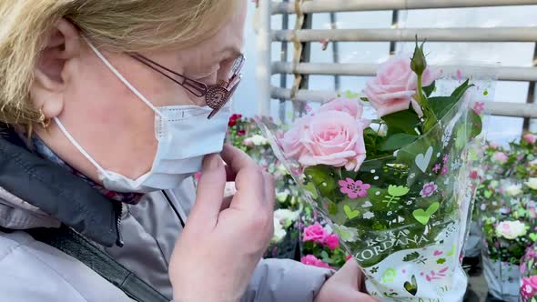 Senior woman in a medical mask sniffs pink potted roses. Flowers as a gift