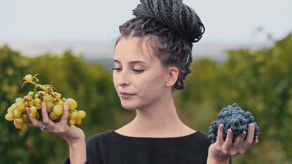 Young Woman with Dreadlocks Holding White and Black Grapes in Her Hands and Bites Off a Few Pieces