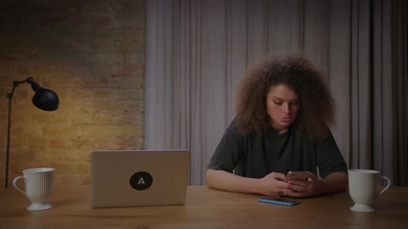 Young Adult Woman Using Mobile Phone for Texting with Friends Sitting at Table at Home Alone