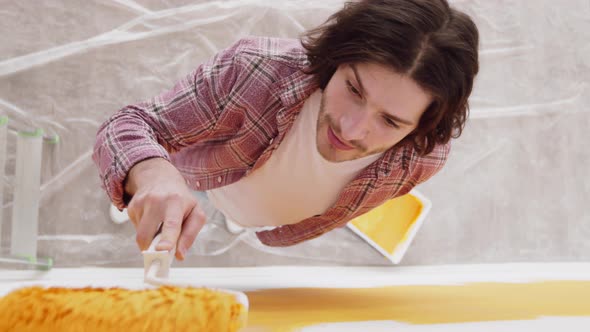 High Angle View on Young Man Painting Wall with Yellow Paint on Roller Smiling at Camera