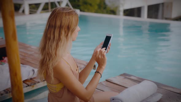 A Blonde Woman Uses Her Mobile Phone While Lounging in the Sun By the Pool on Vacation