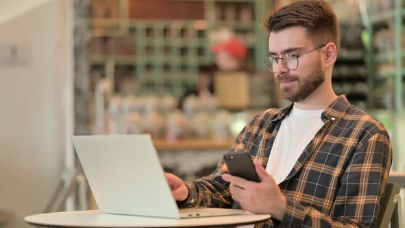 Young Man Using Smartphone and Laptop in Cafe