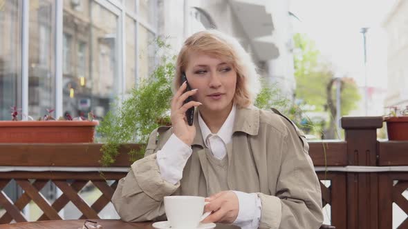 Woman Talking on a Cell Phone with a Cup of Coffee at a Table on the Terrace