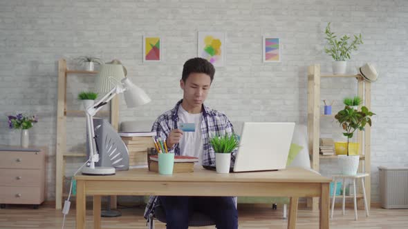 Young Asian Man in a Shirt Enters Data with a Bank Card on a Laptop