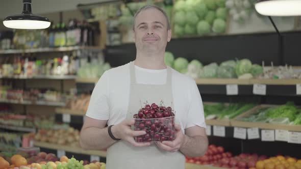 Portrait of Positive Caucasian Trader Posing with Basket of Sweet Cherry in Grocery. Adult Smiling