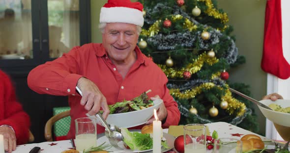 Happy caucasian senior man in santa hat serving himself food, at christmas dinner table with friends