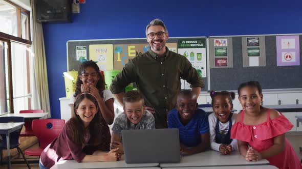 Portrait of happy diverse male teacher and group of schoolchildren looking at laptop