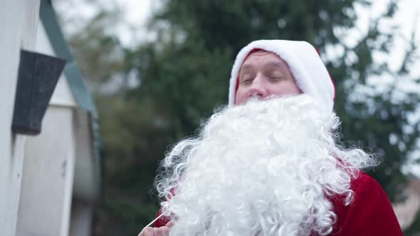 Portrait of Man in Red Costume with White Beard Ringing Doorbell Standing Outdoors