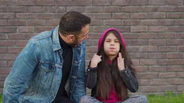 Dad and Daughter Are Sitting, Having Fun and Chewing Bubble Gum Against the Backdrop of Grass and a