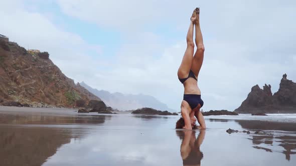 Sports Girl Standing on Her Head on the Ocean Moves Her Legs in the Air