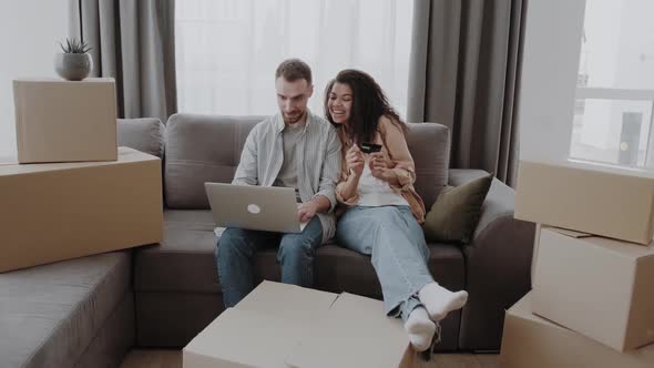 Happy Couple Beween a Heap of Boxes in Living Room Sitting at the Couch Use a Laptop and Credit Card
