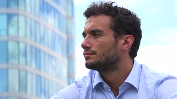 A Young Handsome Businessman Looks Around Thoughtfully - Closeup - an Office Building in Background