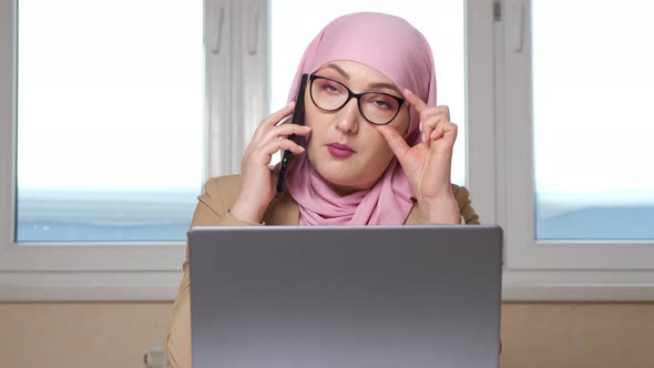 Muslim Woman in Pink Headscarf and Glasses Talking on the Phone While Sitting at the Table with