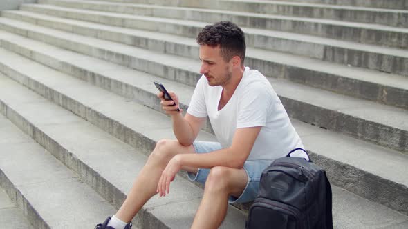 Handsome Young Man Sitting on the Steps of the Stairs in the City and Flipping the News Feed on His