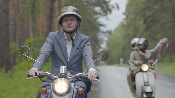 Positive Confident Man Riding on Scooter While Couple of Friends Driving at the Background Shooting