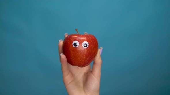 Red Apple with Eyes in a Woman Hand. Apple Jumps Into the Frame and Looks Around on a Blue