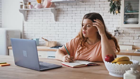 Concentrated Plus Size Female Student Watching Webinar on Laptop and Making Notes Sitting at Kitchen