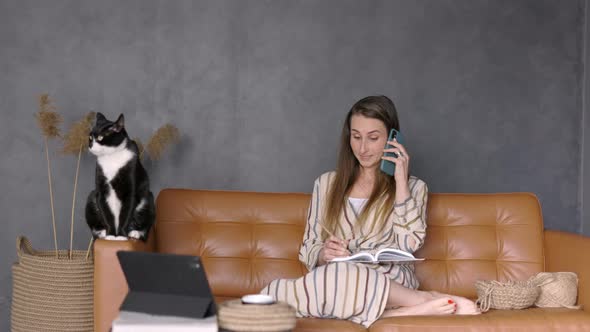 Young Woman having a Phone Call, taking Notes at Home sitting on a Couch with her Cat, Copy Space