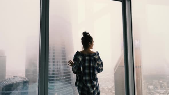 Attractive Girl Stands at the Window Against the Background of the City of Skyscrapers