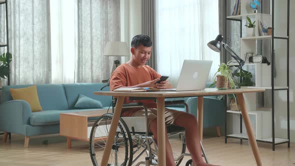 Young Asian Boy Sitting In A Wheelchair While Using Laptop Computer And Use Mobile Phone At Home