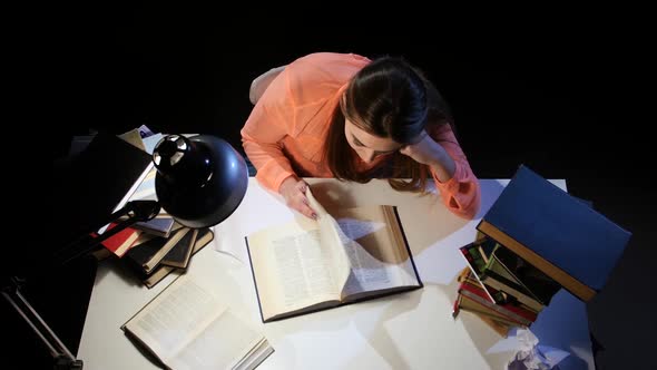 Girl Leafing Through a Book and Falling Asleep at the Table. Black Background. View From Above