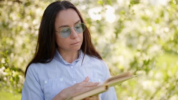 Funny Book Lover Woman with Round Glasses is Reading Old Book in Garden in Spring Day Portrait