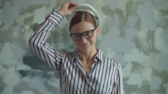 Young 30s Woman in Glasses Putting on White Hardhat Looking at Camera. Smiling Female Foreman Is