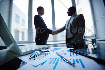 touchpad and financial documents at workplace with businessmen handshaking on background