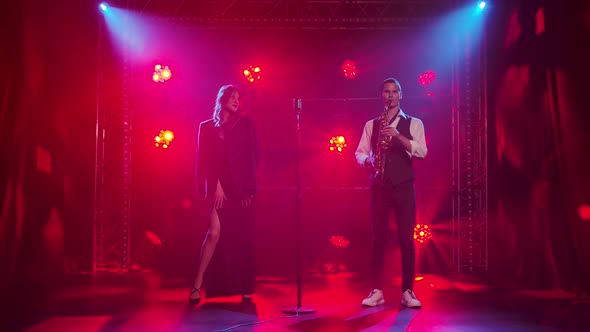 A Young Stylish Guy Plays the Saxophone on Stage in the Rays of Red Spotlights. In the Background