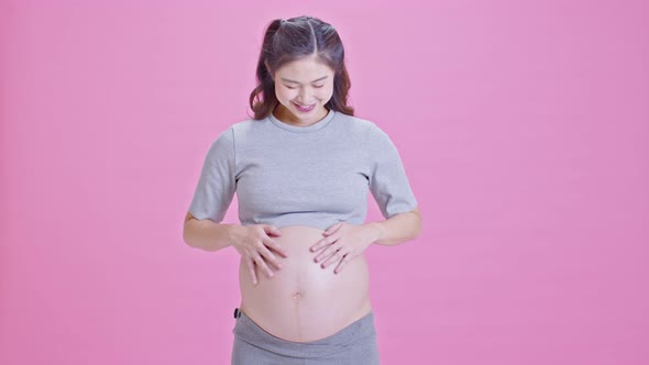 Pregnant Woman standing smile stroking big belly showing thumbs up sign isolated on pink
