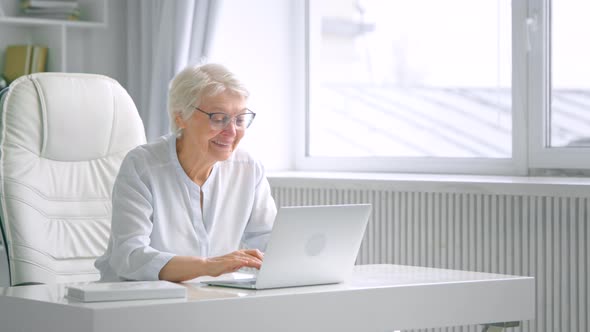 Elegant old lady in glasses types on computer communicating with friend