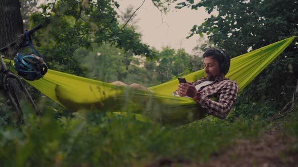 Man on Bicycle Trip at Camping By Lake is Relaxing in Green Hammock While Listening to Music
