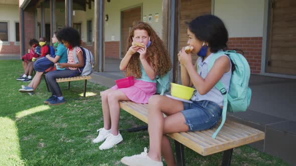 Two girls eating lunch from tiffin box while sitting on bench in the park at school