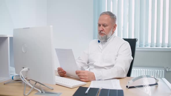 Grayhaired Male Doctor Sits at Workplace and Communicates with Colleagues Via Video Call Remote