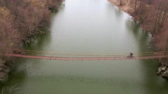 Aerial Slowmotion Shot of Young Sport Man Rides Bicycle on Suspension Bridge Over Small River at