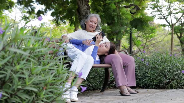 An elderly mother and Asian daughter lounging in the park enjoying their vacation.