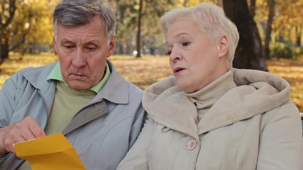 Mature Couple Grandparents Sit on Bench in Autumn Park Receive Letter Open Envelope Correspondence