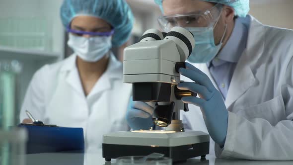 Researcher Viewing Sample Under Microscope, Assistant Taking Notes, Laboratory