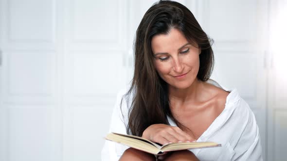 Portrait of Smiling Pretty Woman Reading Book and Turning Page Sitting on Bed in Bedroom