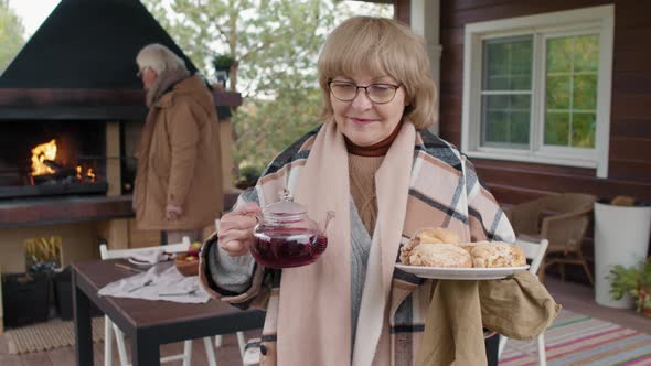 Aged Woman Holding Teapot and Pie on Terrace