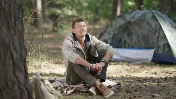 Satisfied Caucasian Man Sitting in Forest at Tent Smiling Looking Around
