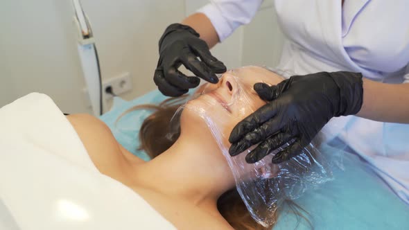 The Cosmetologist Applies Mask and Wrapping Film on Top of the Patient's Fair Skin