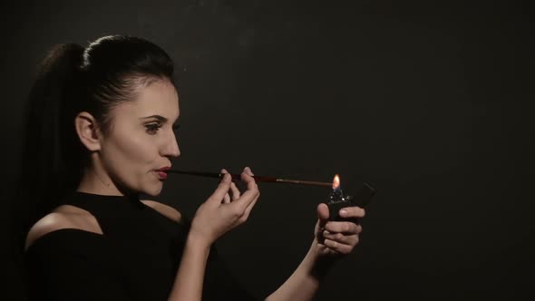 Beautiful Woman Lights a Cigarette in a Mouthpiece on a Black Background