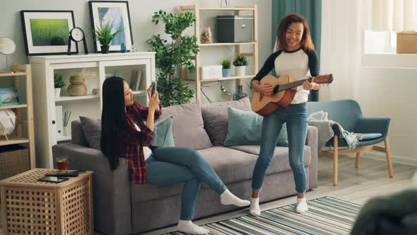 Cute African American Woman Is Playing the Guitar at Home While Her Asian Friend Is Recording Video