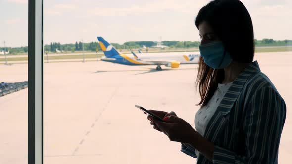 Portrait of Woman in Mask, Uses Mobile, Stands Against Panoramic Window with Runway and Big Plane