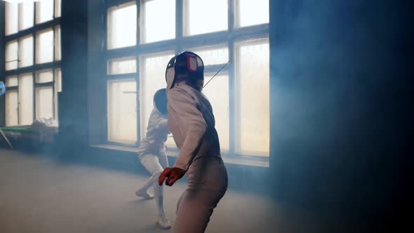 Two Young Women Fencers Having a Training Duel in the Smoky Studio