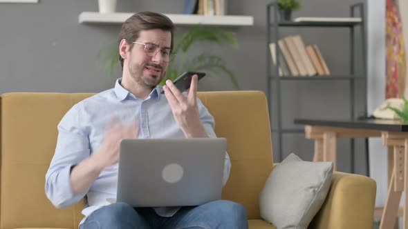 Angry Young Man with Laptop Talking on Smartphone on Sofa
