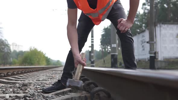 A railroad worker in a hard hat is repairing a railroad by hammering a spire with a sledgehammer.