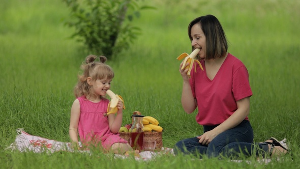 Family Weekend at Picnic. Daughter Child Girl with Mother on Grass Meadow Eating Bananas, Having Fun