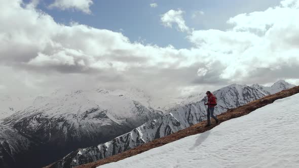 Aerial View Young Man in Sunglasses with a Backpack Goes Down the Mountain on the Crest on the